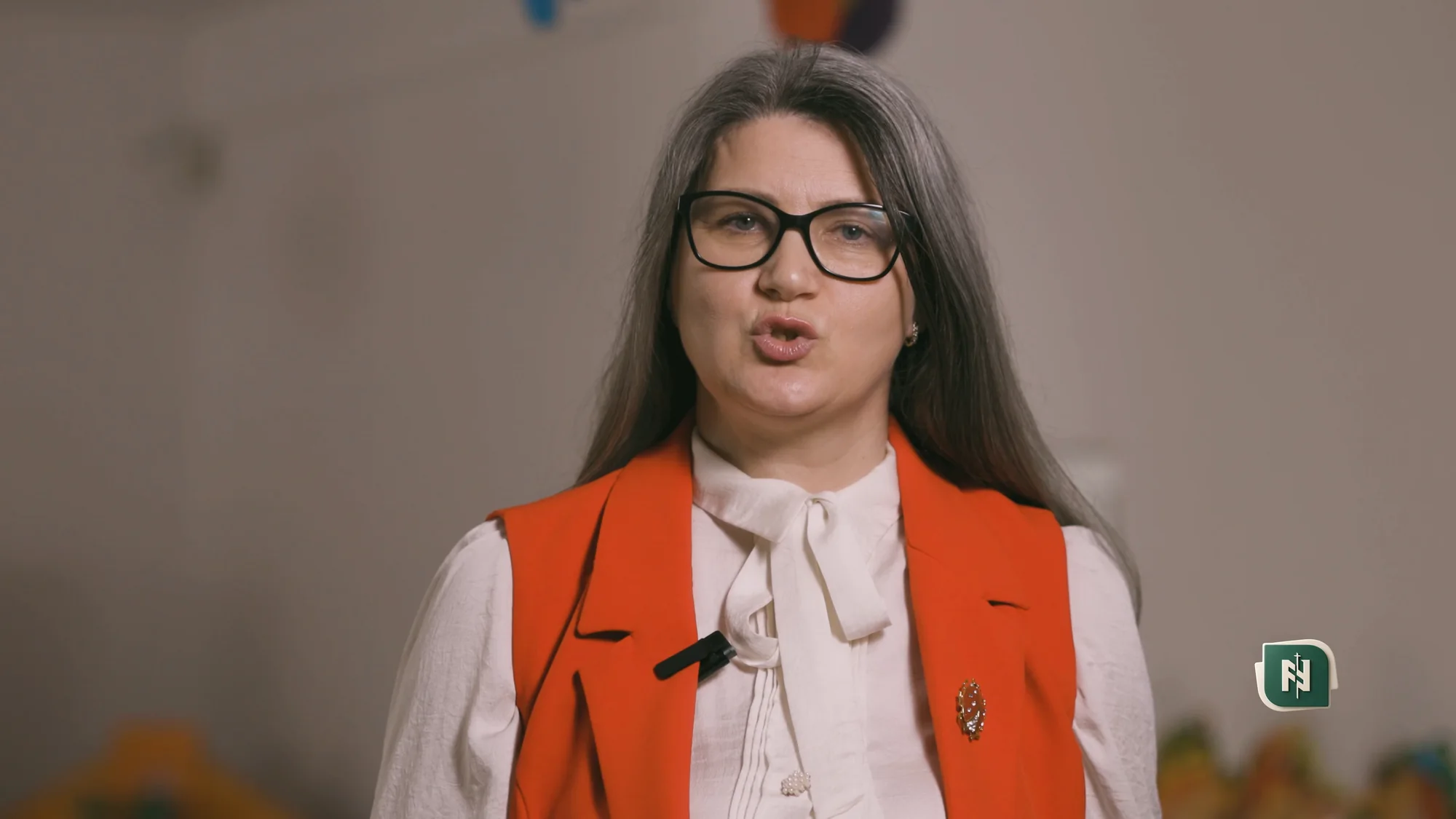 O profesoară a liceului, cu ochelari și vestă portocalie, filmată orizontal.