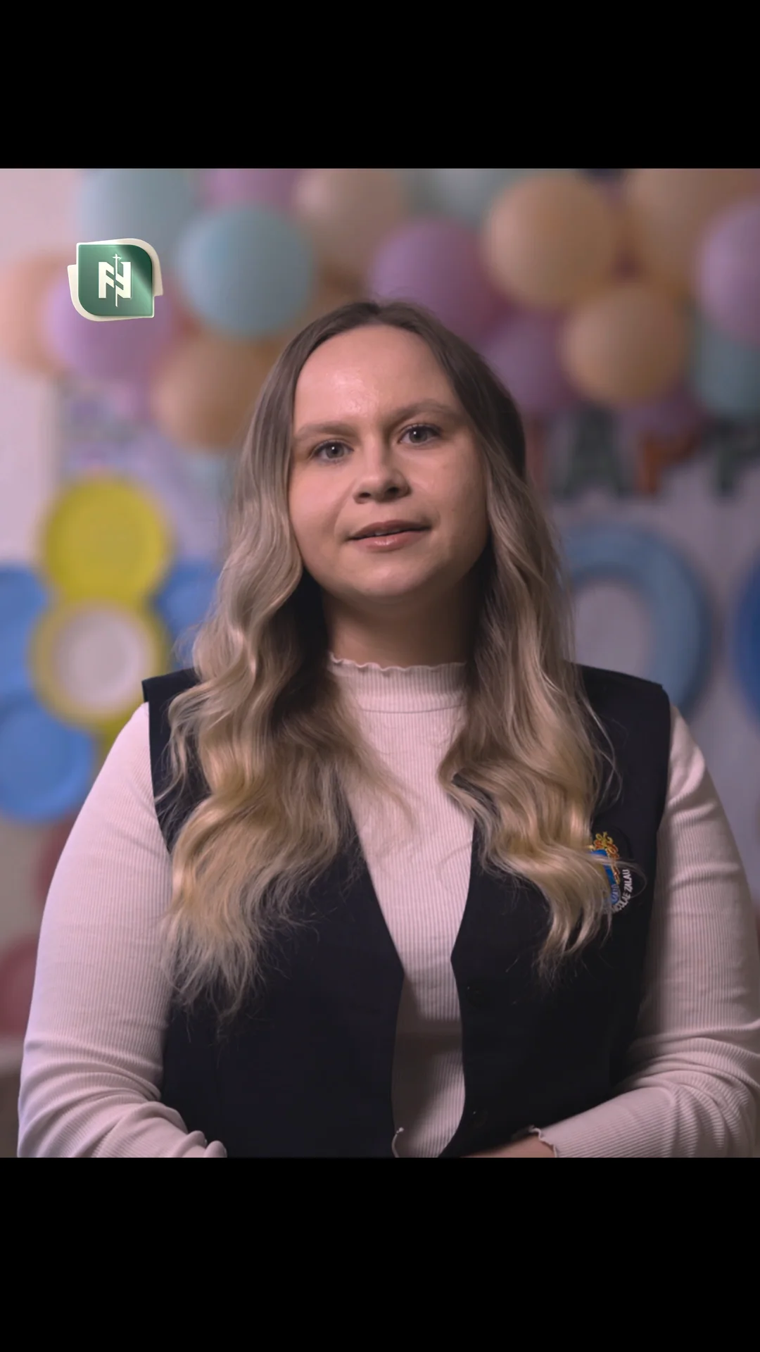 O învățătoare, filmată pe un fundal cu baloane colorate.